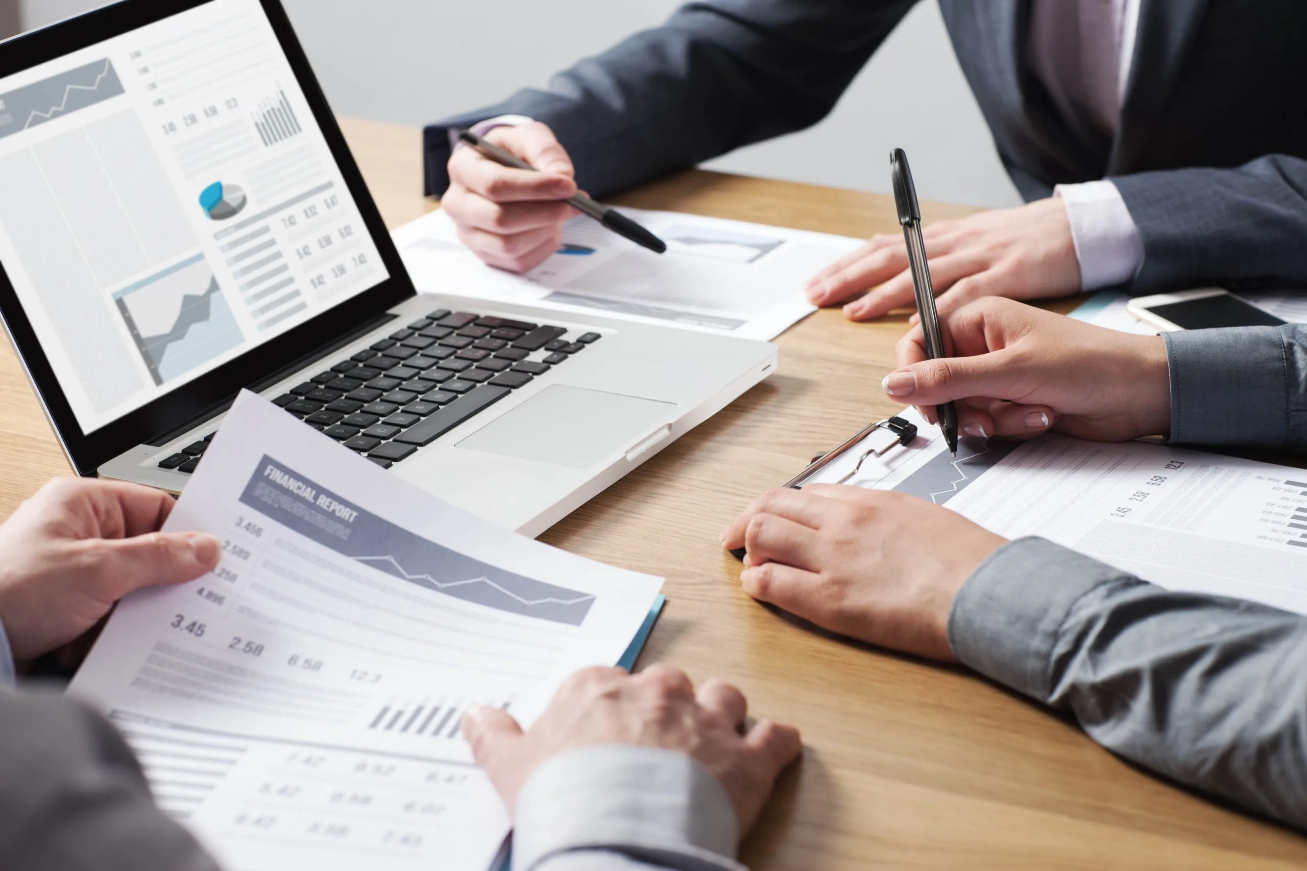 Three people reviewing financial report printouts and laptop charts at a table