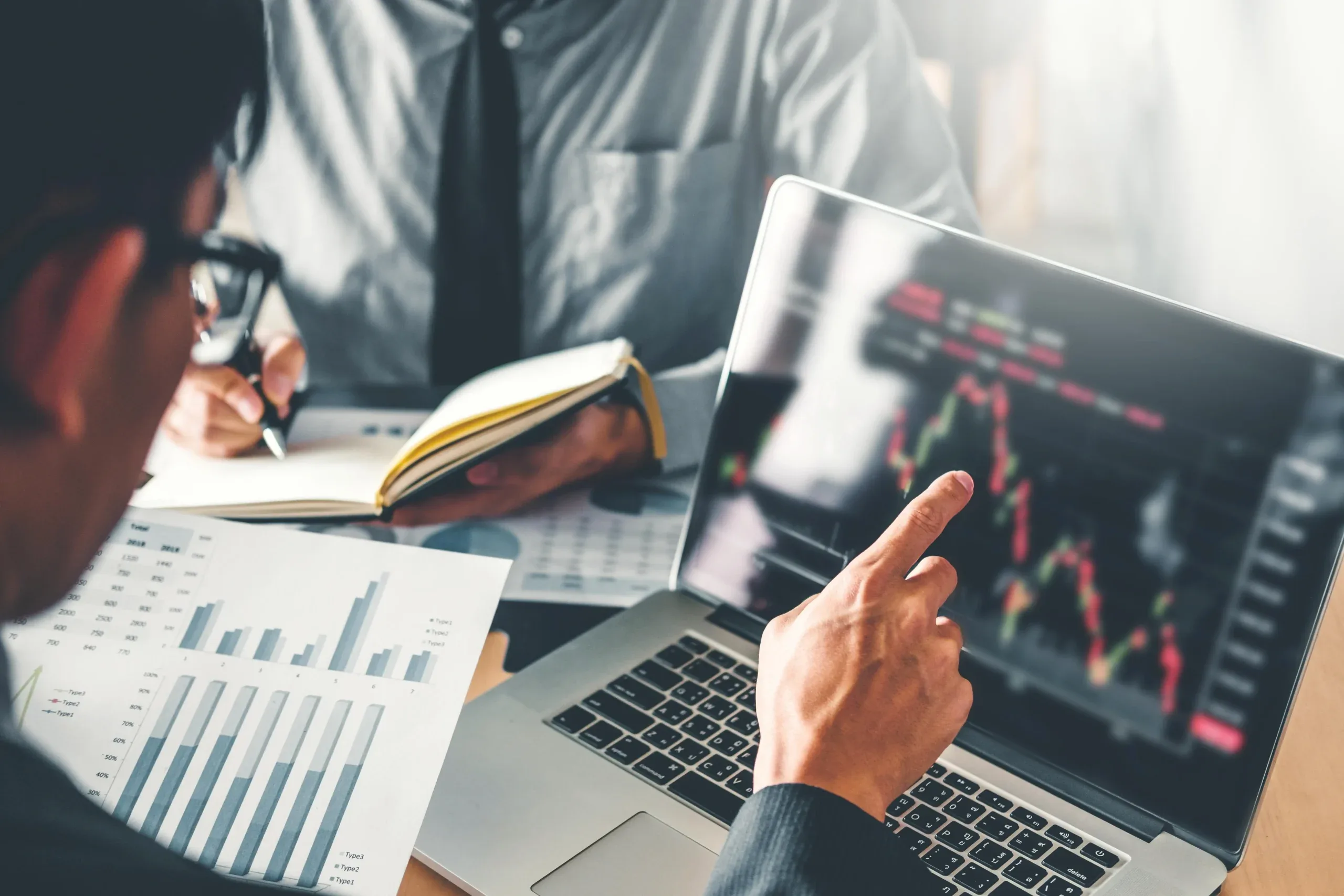 Two investors comparing stock charts on a laptop with printed bar graphs