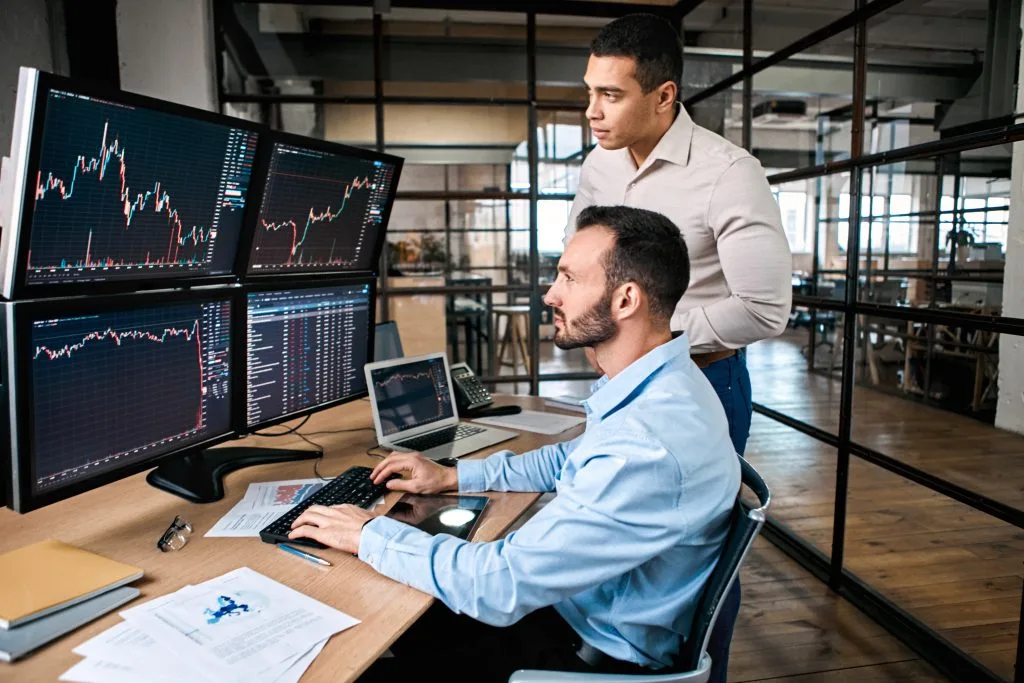 Two traders reviewing candlestick charts on multiple monitors in an office
